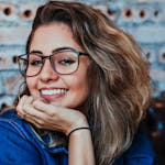 A young woman wearing glasses and denim, smiling in an indoor setting.